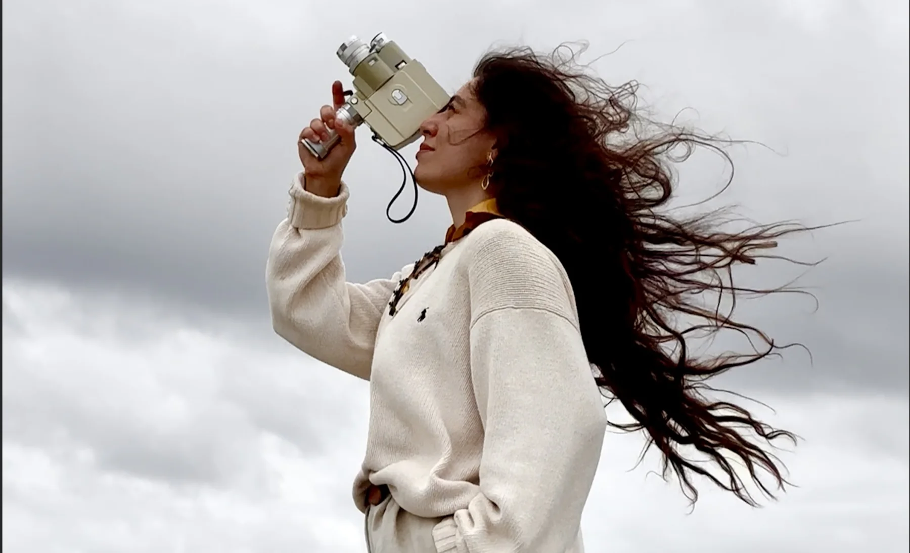 Carmen Sanchez looking through a cine camera as her hair blows beneath an overcast sky.